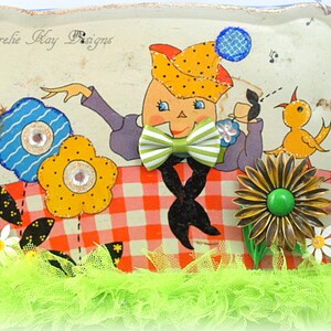 May include: A vintage tin tray with a whimsical illustration of a cartoon character wearing a hat and bow tie, holding a cup. The character is surrounded by flowers and a bird. The tray has a red and white checkered pattern and is decorated with green, blue, and yellow accents. The text "Lorelie Kay Designs" is visible in the top left corner.