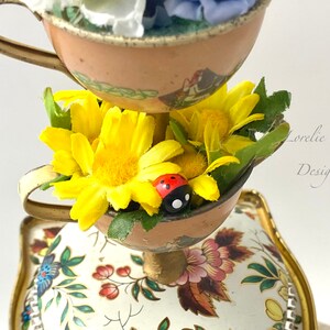 May include: A tiered teacup centerpiece with a white background and floral designs. The top tier has a pink teacup with white and blue flowers. The middle tier has a brown teacup with yellow flowers and a red ladybug. The bottom tier has a white teacup with a floral design.