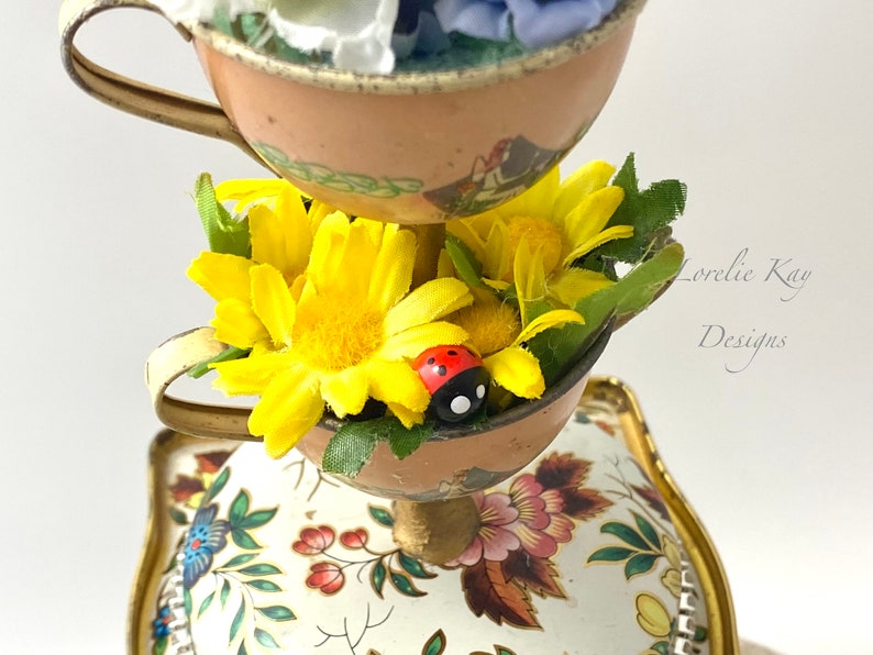May include: A tiered teacup centerpiece with a white background and floral designs. The top tier has a pink teacup with white and blue flowers. The middle tier has a brown teacup with yellow flowers and a red ladybug. The bottom tier has a white teacup with a floral design.