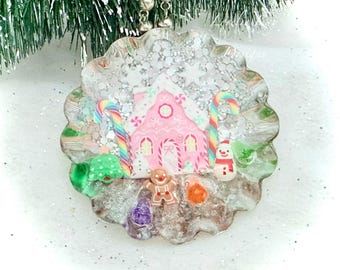 May include: A round, scalloped-edge clear resin ornament filled with silver glitter. Inside, a pink gingerbread house is flanked by two rainbow candy canes, a small green Christmas tree, a snowman, and a gingerbread man figure.