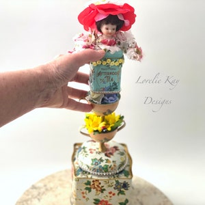 May include: A whimsical tower made of vintage tea tins, porcelain cups, and flowers. The top features a porcelain doll wearing a red flower hat. The tower is decorated with rhinestones and beads. The text "Lorelie Kay Designs" is visible in the background.