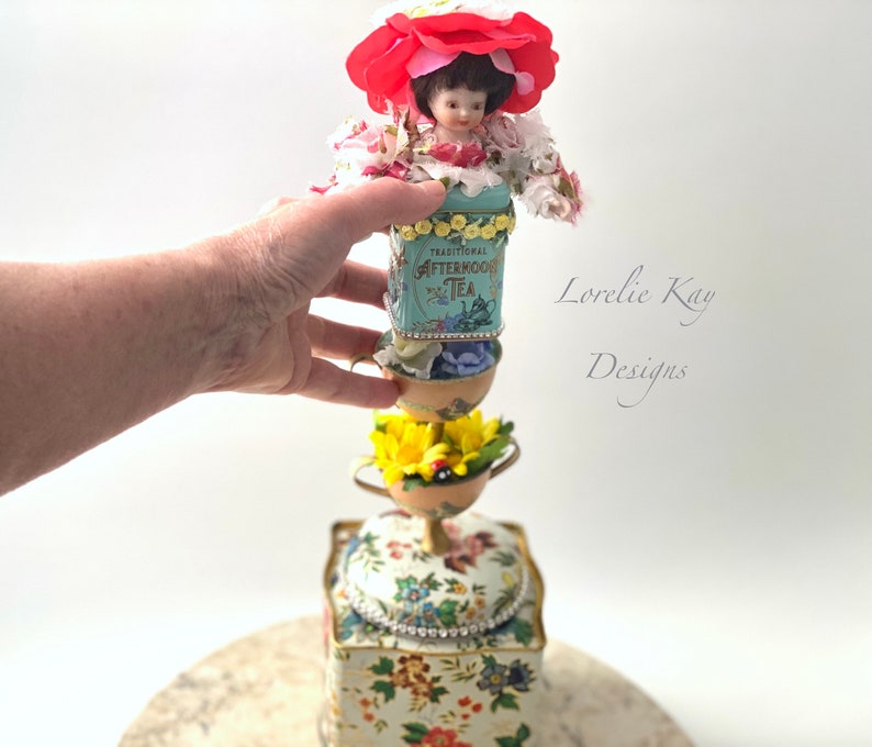 May include: A whimsical tower made of vintage tea tins, porcelain cups, and flowers. The top features a porcelain doll wearing a red flower hat. The tower is decorated with rhinestones and beads. The text "Lorelie Kay Designs" is visible in the background.