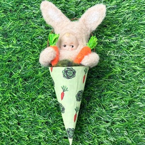 May include: A felt bunny wearing a baby doll costume holds two carrots in a green and white cone-shaped container with a carrot and cabbage print. The cone is sitting on a green grass surface.