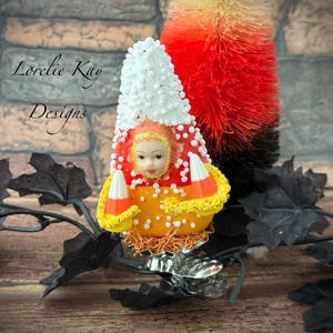 May include: A novelty Halloween ornament in the shape of a candy corn person with a doll face. The figure is orange, yellow, red, and white, with a decorative base and a small, colourful tree in the background. The text "Lorelie Kay Designs" is visible.