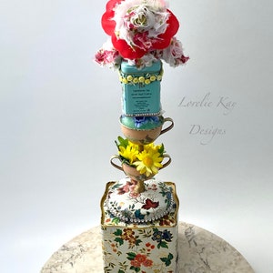 May include: A tiered tower made of teacups and a floral patterned box. The top of the tower is decorated with a red and white fabric flower. The box has a gold trim and rhinestones. The text "Lovelie Kay Designs" is visible on the right side of the image.