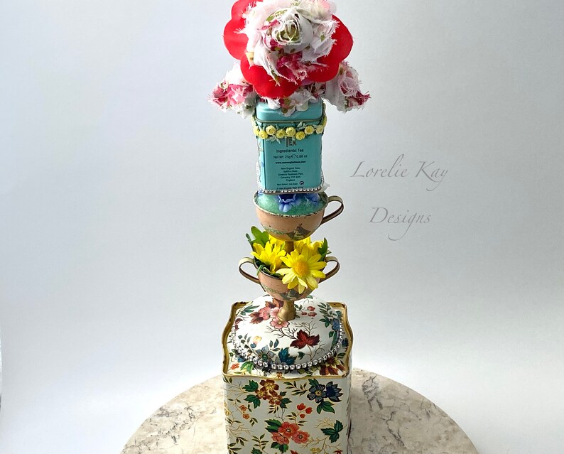 May include: A tiered tower made of teacups and a floral patterned box. The top of the tower is decorated with a red and white fabric flower. The box has a gold trim and rhinestones. The text "Lovelie Kay Designs" is visible on the right side of the image.