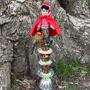 May include: A red hooded doll with a basket of fruit stands on a tower of upcycled teacups and saucers. The tower is decorated with flowers and mushrooms. The doll is standing in front of a tree trunk.