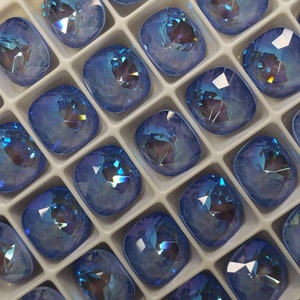 May include: A close-up view of a tray filled with light blue, faceted, cushion-shaped rhinestones. The rhinestones are arranged in rows and columns, and they have a shimmering, iridescent finish.