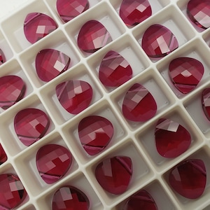 May include: A close-up view of a white plastic tray filled with small, faceted, teardrop-shaped, pink glass rhinestones.