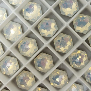 May include: A close-up view of a tray of iridescent white, faceted, hexagonal rhinestones. The rhinestones are arranged in rows in a white plastic tray with a brown cardboard border.