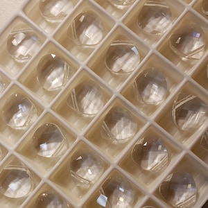 May include: A close-up view of a white plastic tray filled with clear, faceted, heart-shaped gemstones. The tray is filled with rows of gemstones, each one reflecting light.