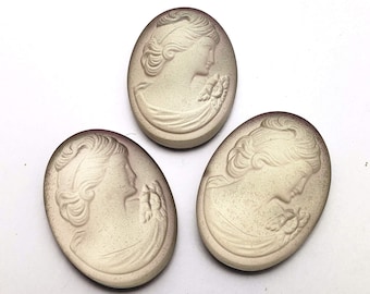 May include: Three white cameo-style ceramic buttons with a raised design of a woman's profile. The buttons have a slightly oval shape and a subtle purple hue around the edges.
