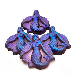 Puede incluir: Cuatro figuritas de sirena iridiscentes de color morado y azul, cada una sentada en una concha, están dispuestas en forma de pirámide.