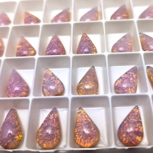 May include: A tray of teardrop-shaped gemstones in shades of purple, orange, and gold. The gems are arranged in a grid of white compartments, showcasing their unique patterns and colors. The gemstones have a polished, reflective surface.