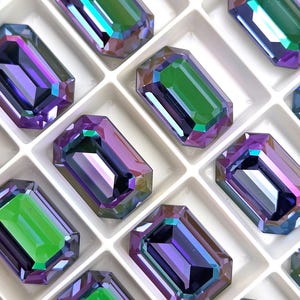 Op de afbeelding: Close-up van verschillende iriserende, smaragdgeslepen kristallen in een witte tray. De kristallen hebben een mix van paarse, groene en blauwe tinten, met een rechthoekige vorm en facetranden. De kristallen zijn gerangschikt in een rasterpatroon.