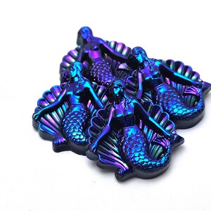 May include: A collection of three iridescent mermaid figurines, each featuring a metallic finish that shifts between deep blue, purple, and green tones. Each mermaid is depicted sitting in front of a large, fan-shaped seashell structure.