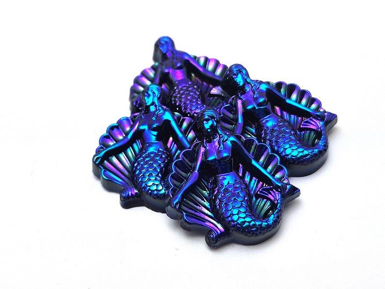 May include: A collection of three iridescent mermaid figurines, each featuring a metallic finish that shifts between deep blue, purple, and green tones. Each mermaid is depicted sitting in front of a large, fan-shaped seashell structure.