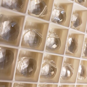 May include: A close-up of a white plastic container filled with clear glass crystal prisms. The prisms are arranged in rows and columns, and they reflect light in a variety of colors.