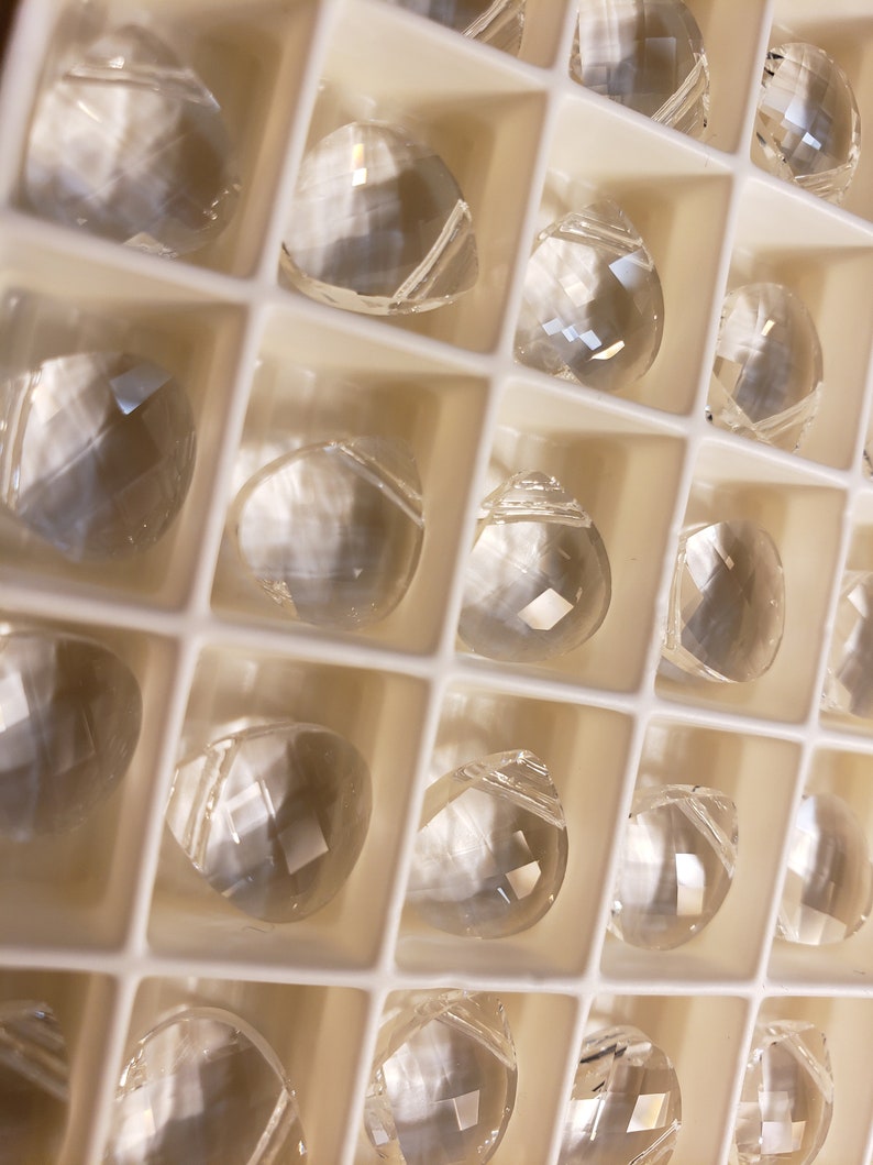 May include: A close-up of a white plastic container filled with clear glass crystal prisms. The prisms are arranged in rows and columns, and they reflect light in a variety of colors.