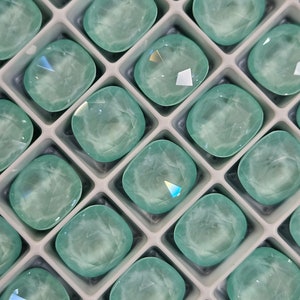 May include: A close-up view of a tray of light blue, faceted, cushion-shaped gemstones. The gemstones are arranged in a grid pattern and are set in a white tray.
