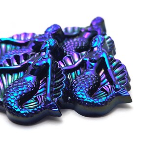 May include: A collection of iridescent mermaid-shaped figures with a metallic finish that shifts between deep blue, purple, and green tones. Each figure depicts a mermaid resting against a large, textured seashell, showcasing detailed scales and hair.