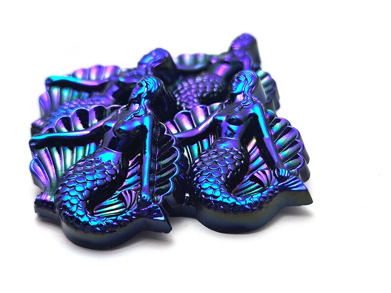May include: A collection of iridescent mermaid-shaped figures with a metallic finish that shifts between deep blue, purple, and green tones. Each figure depicts a mermaid resting against a large, textured seashell, showcasing detailed scales and hair.