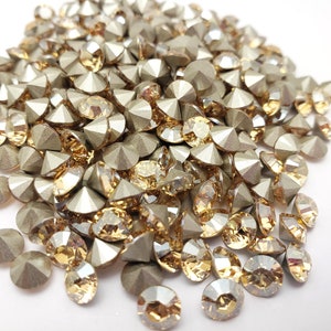 May include: A pile of gold and clear glass rhinestones with a pointed back. The rhinestones are scattered on a white surface.