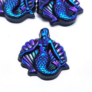May include: Three iridescent mermaid cabochons featuring a metallic finish with shifting blue, purple, and green hues. Each piece depicts a mermaid figure centered in front of a scalloped seashell, showcasing detailed scales and defined facial features.