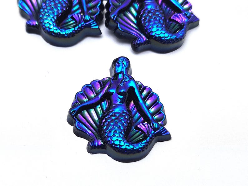 May include: Three iridescent mermaid cabochons featuring a metallic finish with shifting blue, purple, and green hues. Each piece depicts a mermaid figure centered in front of a scalloped seashell, showcasing detailed scales and defined facial features.