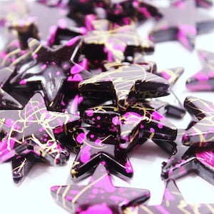 May include: A group of black star-shaped resin beads with pink and gold accents. The beads are scattered on a white surface.