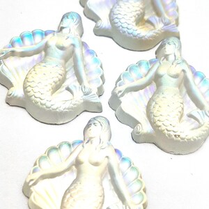 May include: Four iridescent white mermaid figurines, each sitting in a shell with a pearl-like finish. The mermaids have long flowing hair and are in a sitting position.