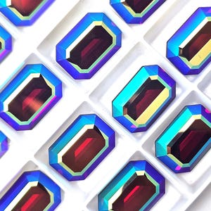 May include: Close-up of several iridescent, emerald-cut gemstones. Each gem has a deep red center, framed by a gold, teal, and blue border. The gems are arranged in a white tray, creating a visually appealing pattern.