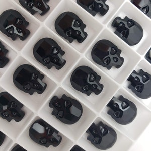 May include: A white plastic tray filled with black skull-shaped glass rhinestones. The rhinestones are arranged in rows and columns.