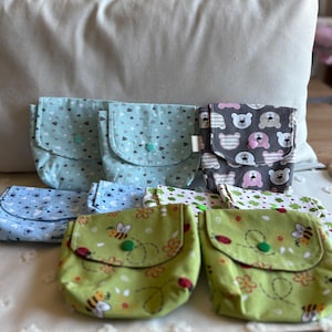 Może przedstawiać: Kolekcja ręcznie robionych materiałowych torebek w różnych wzorach i kolorach. Torebki mają zapięcia na klapę z guzikami. Wzory obejmują jasnoniebieski z sercami, zielony z pszczołami i szary z twarzami niedźwiedzi.