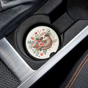 May include: A white car coaster with a floral design and a sloth holding a bouquet of flowers. The coaster is in a car cup holder.