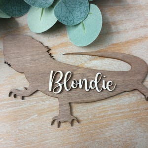 Puede incluir: Un letrero de madera marrón claro con forma de dragón barbudo. El nombre "Blondie" está escrito en blanco con una fuente cursiva en el cuerpo del lagarto. El letrero tiene patas con garras detalladas y una cola larga.