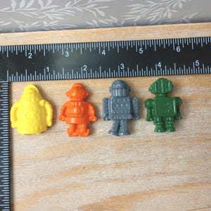 May include: Four colorful robot figurines in yellow, orange, gray, and green are arranged on a wooden surface. A black ruler with inch measurements is placed next to the robots. The robots are small, plastic toys.
