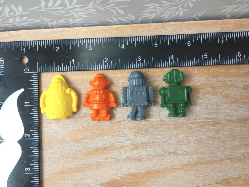 May include: Four colorful robot figurines in yellow, orange, gray, and green are arranged on a wooden surface. A black ruler with inch measurements is placed next to the robots. The robots are small, plastic toys.