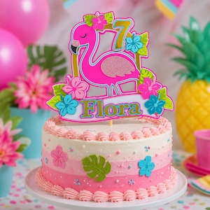 May include: A round cake with pink and white ombre frosting, decorated with green leaf and flower cutouts. A cake topper features a bright pink flamingo, a gold number 7, and a white banner with the name Flora in gold lettering.