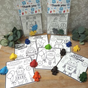 May include: A collection of robot-themed birthday party favors. The favors include coloring pages with robot illustrations and the text "Happy Birthday", "Robbie", and "Thank you". Small, colorful robot-shaped crayons are scattered around the pages.