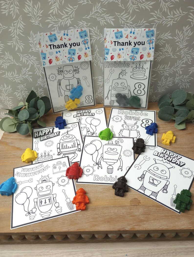 May include: A collection of robot-themed birthday party favors. The favors include coloring pages with robot illustrations and the text "Happy Birthday", "Robbie", and "Thank you". Small, colorful robot-shaped crayons are scattered around the pages.
