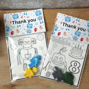 May include: Two clear plastic bags with a white top featuring the text "Thank you" and robot illustrations. The bags contain coloring pages with robot designs and small robot-shaped crayons. One page has the name "Robbie" and the number "8".