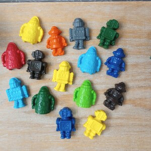 May include: A collection of colorful, plastic robot figurines in various colors, including yellow, orange, red, gray, green, blue, and black. The robots are arranged on a light-colored wooden surface.