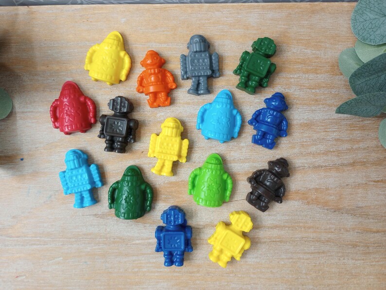 May include: A collection of colorful, plastic robot figurines in various colors, including yellow, orange, red, gray, green, blue, and black. The robots are arranged on a light-colored wooden surface.