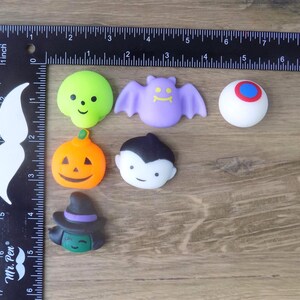 May include: A collection of six small Halloween-themed silicone charms including a lime green skull, a purple bat, a white eyeball with a blue and red iris, an orange pumpkin, a vampire, and a witch, measured by an L-shaped ruler up to 6 inches.