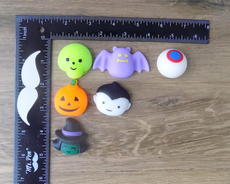 May include: A collection of six small Halloween-themed silicone charms including a lime green skull, a purple bat, a white eyeball with a blue and red iris, an orange pumpkin, a vampire, and a witch, measured by an L-shaped ruler up to 6 inches.