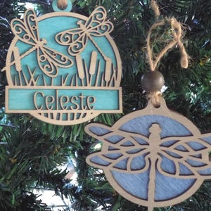 May include: Two wooden dragonfly ornaments with a light blue background. One ornament has the name "Celeste" on it. The ornaments are hanging on a Christmas tree. The dragonfly designs are cut out of the wood.