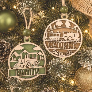 May include: Two circular wooden ornaments featuring laser-cut train designs. One ornament is natural wood with the name GEORGE, and the other is green with the name DANNY. Both hang from jute twine with a green bead and depict a train on tracks.