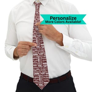 May include: A burgundy tie with the word "Hubby" printed repeatedly in white. The tie is being held by a person wearing a white dress shirt. The image includes the text "Personalize More Colors Available!"