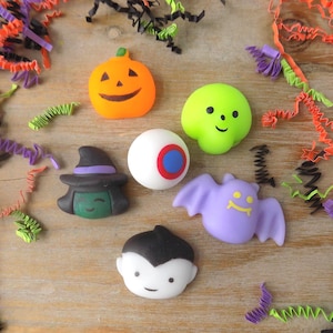 May include: A collection of six small Halloween-themed figurines including an orange jack-o'-lantern, a lime green skull, a white eyeball with a blue iris, a green-faced witch with a black hat, a purple bat, and a vampire with black hair.